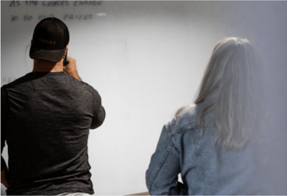 View of the backs of a man with a backwards hat and woman with jean jacket stand in front of a whiteboard