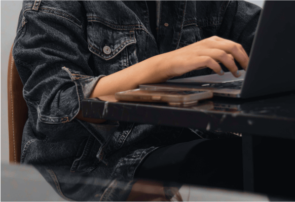 Close up of a person in a jean jacket working on a laptop and smartphone on their desk