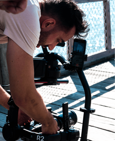 A man in a white tshirt leaning over with a camera at a videoshoot